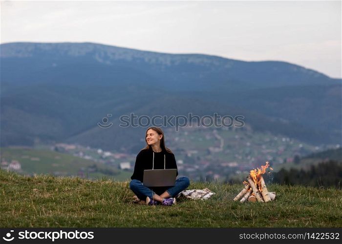young woman freelancer with laptop in the mountains in the evening. Tourist girl sitting near campfire. young woman freelancer with laptop in the mountains in the evening. Tourist girl sitting near campfire.