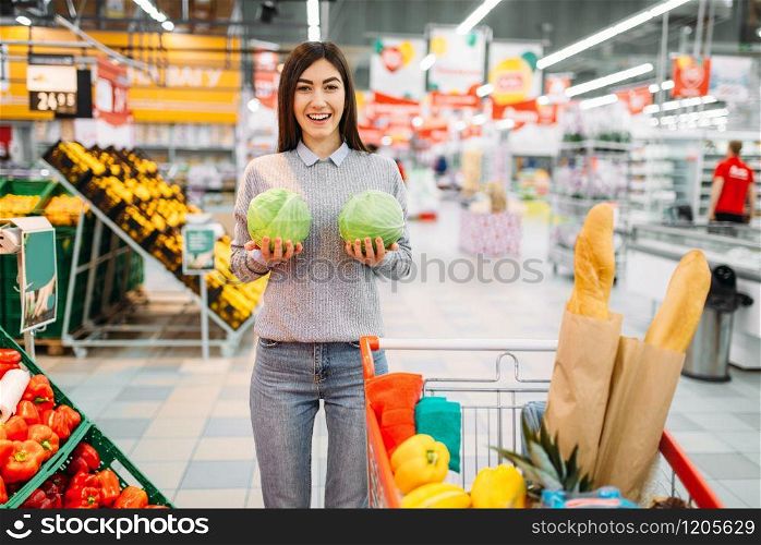 Young woman funs with cabbage swings in a supermarket, family shopping. Customer in shop, buyer in market