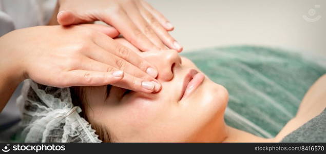 Young woman getting facial massage with closed eyes by beautician in beauty salon. Young woman getting facial massage