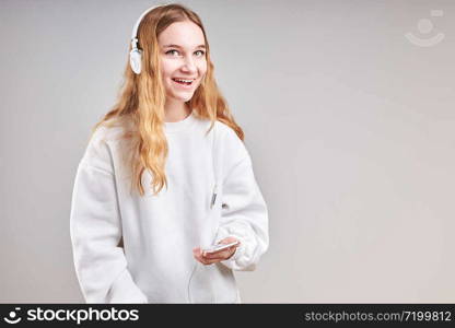 Young woman girl listening to music streaming content having fun watching video enjoying video chat talking with friends making gestures faces using smartphone earphones headphones standing over plain grey background