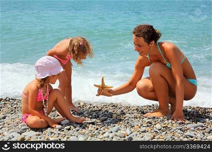 young woman gives starfish to two little girls on stony beach