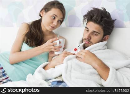 Young woman giving coffee mug to man with thermometer in mouth