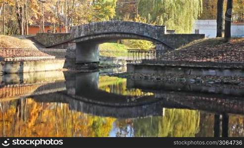 young woman goes down to lake near small stone bridge in beautiful autumn city park