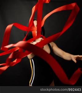 young woman gymnast of Caucasian appearance with black hair spins red satin ribbons, gymnastic exercises on black background, selective focus