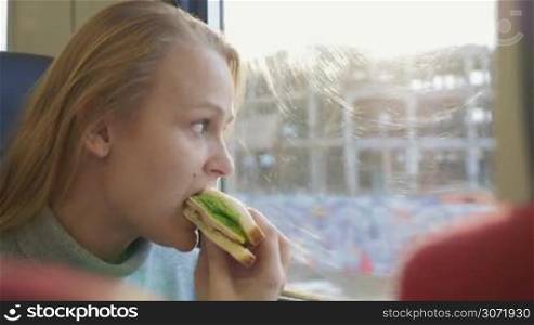 Young woman having a bite in the train while looking out the window. She eating sandwich with fresh salad and drinking water