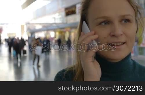 Young woman having a phone talk in the station or airport hall. Defocused crowd of people in background