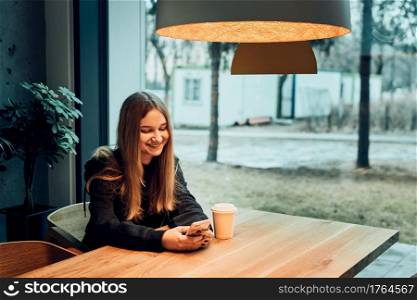 Young woman having a video call, talking remotely, drinking coffee, sitting in a cafe. Girl relaxing in cafe using mobile phone