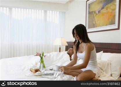 Young woman having breakfast in bed with a mid adult man sleeping beside her