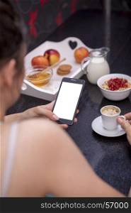 Young woman having breakfast while using smartphone with blank screen