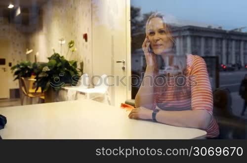 Young woman having excited conversation on the phone sitting in cafe in the evening. City reflecting in the glass