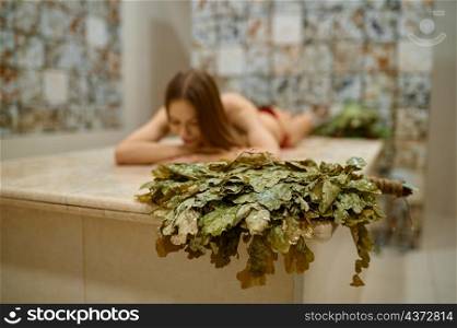 Young woman having rest in sauna alone. Relaxation and spa procedure in bathhouse. Focus on broom. Woman rest in bathhouse, focus on broom
