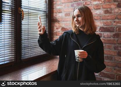 Young woman having video call, talking remotely, taking selfie photo holding smartphone. Girl drinking coffee relaxing while taking break in office standing at window
