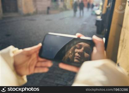 Young woman having video call, talking remotely, taking selfie photo holding smartphone. Face reflected in screen of smartphone