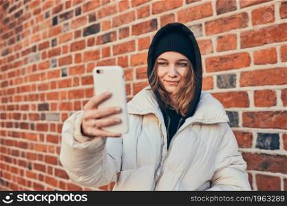 Young woman having video call, talking remotely, taking selfie photo holding smartphone standing at brick wall