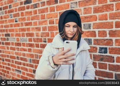 Young woman having video call, talking remotely, taking selfie photo holding smartphone standing at brick wall