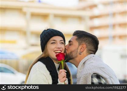 Young woman holding a red rose laughs as she receives a romantic kiss on the cheek by a young man on an out of focus background. Valentine?s concept.. Young woman laughs as she receives a kiss