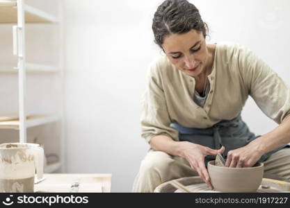 Young woman holding pottery instrument for scraping, smoothing, shaping and sculpting. Lady siting on bench with pottery wheel and making clay pot. Lady siting on bench with pottery wheel and making clay pot