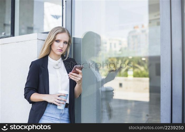 young woman holding takeaway coffee cup mobile phone leaning reflective glass