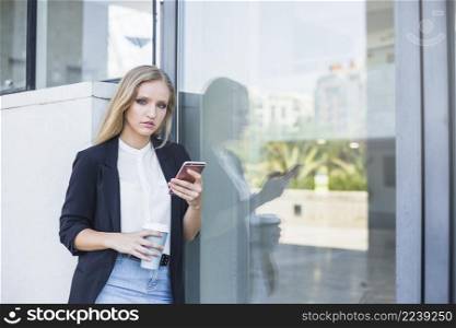 young woman holding takeaway coffee cup mobile phone leaning reflective glass