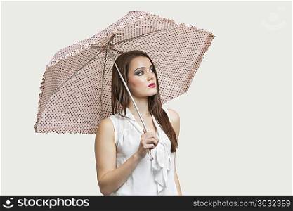 Young woman holding umbrella while looking away over gray background