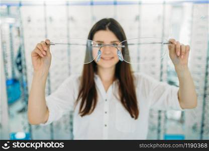 Young woman holds huge decorative glasses, optic store, showcase with spectacles on background. Professional eye care, eyeglasses choice. Woman holds huge decorative glasses, optic store