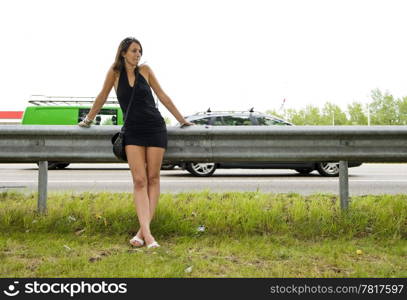 Young woman in a black mini dress leaning on the safety barrier alongside a highway