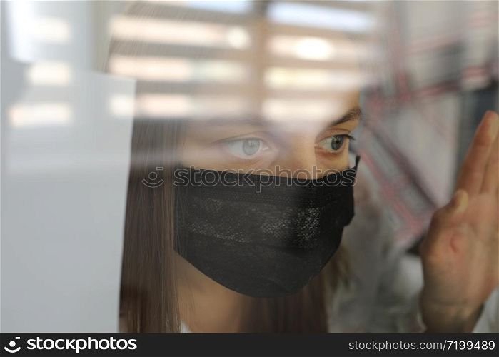 young woman in a medical mask behind a windows glass at home. concept of quarantine of the coronavirus pandemic, covid 19. selective focus.. young woman in a medical mask behind a windows glass at home. concept of quarantine of the coronavirus pandemic, covid 19. selective focus