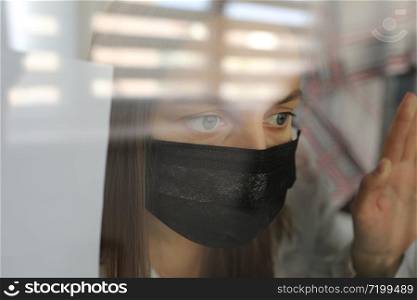 young woman in a medical mask behind a windows glass at home. concept of quarantine of the coronavirus pandemic, covid 19. selective focus.. young woman in a medical mask behind a windows glass at home. concept of quarantine of the coronavirus pandemic, covid 19. selective focus