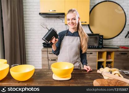 Young woman in apron holds mixer in hands against wooden table with bowls for dough. Sweet cake cooking preparation. Kitchen on background. Woman in apron adds sugar into bowl, dough making