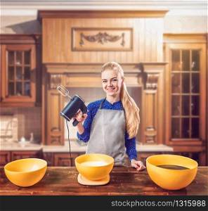 Young woman in apron holds mixer in hands against wooden table with bowls for dough. Sweet cake cooking preparation. Kitchen on background