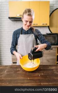 Young woman in apron whip dough in bowl with a mixer. Sweet cake cooking preparation. Kitchen on background