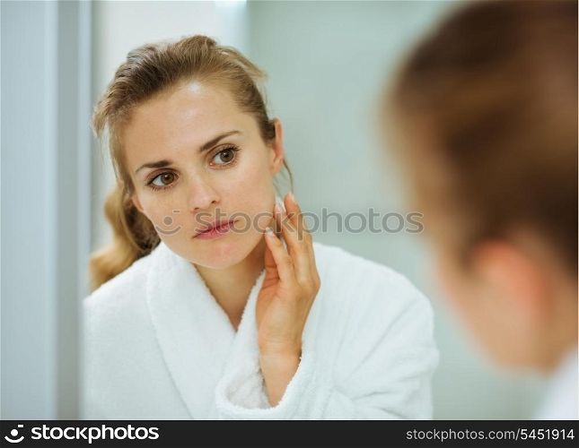 Young woman in bathrobe checking her face in mirror in bathroom