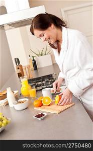Young woman in bathrobe cutting orange for breakfast in kitchen