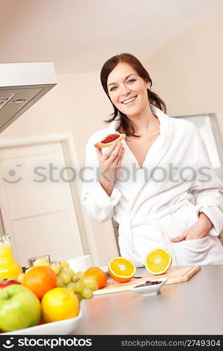 Young woman in bathrobe eating toast for breakfast in kitchen