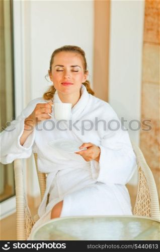 Young woman in bathrobe enjoying cup of coffee on terrace
