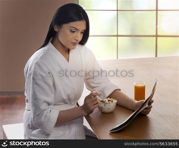Young woman in bathrobe having breakfast while reading newspaper at dining table