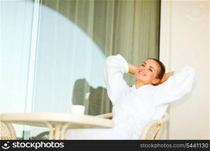 Young woman in bathrobe sitting at table on terrace and relaxing
