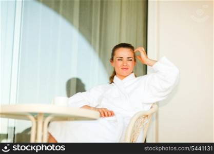 Young woman in bathrobe sitting at table on terrace and thinking