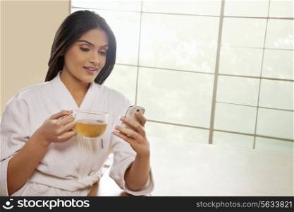 Young woman in bathrobe text messaging while holding tea cup at dining table