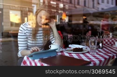 Young woman in cafe using touchpad and eating dessert. View through the glass with reflection of city and people