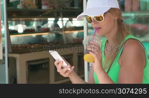 Young woman in cap and sunglasses having cold cocktail and using smart phone. She sitting in the street by the cake shop