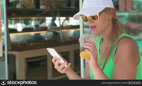 Young woman in cap and sunglasses having cold cocktail and using smart phone. She sitting in the street by the cake shop