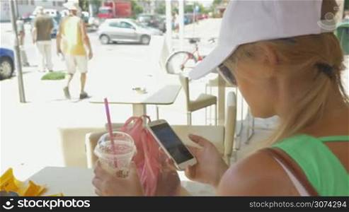 Young woman in cap and sunglasses spending summer day in street cafe. She drinking cold cocktail and chatting in messenger on smart phone