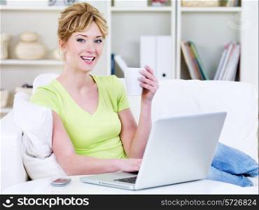 Young woman in casuals with cup of coffee using laptop at home - indoors