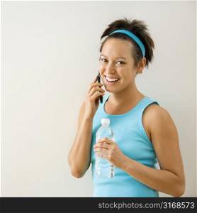 Young woman in fitness clothing holding bottled water talking on cellphone smiling.