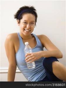 Young woman in fitness outfit sitting on floor with bottled water smiling.