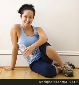 Young woman in fitness outfit sitting on floor with bottled water smiling.