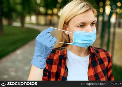 Young woman in gloves puts mask on her face in park, quarantine. Female person walking during the epidemic, health care and protection, pandemic lifestyle. Woman in gloves puts mask on her face, quarantine