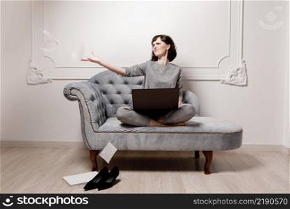 Young woman in gray suit using laptop and having difficulty while working on project and scattering sheets of paper in middle and sitting on sofa in living room. mock up. selective focus.. Young woman in gray suit using laptop and having difficulty while working on project and scattering sheets of paper in middle and sitting on sofa in living room. mock up. selective focus