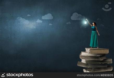 Young woman in green dress with lantern standing on pile of books. Lost in darkness